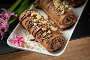 Chocolate roll with chocolate cream with nuts. Chocolate cakes on a white plate, decorated with flowers on a black background.
