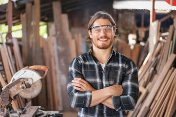 Young male carpenter wear safety glasses smiling with crossed arms working at the carpentry workshop