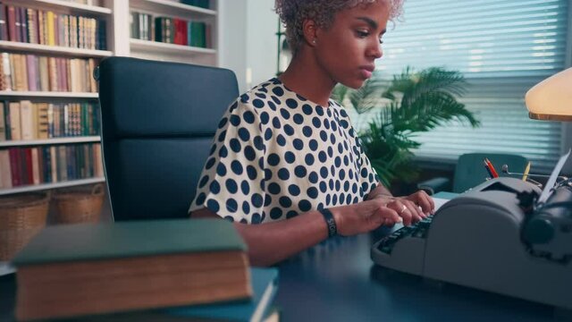 Focused dark skinned female author using old typewriter creates interesting story. Young beautiful and stylish biracial woman with serious expression is typing while sitting in her modern home office.