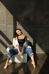 Pretty young woman with big breast posing on white chair in studio, steel wall behind, wearing jeans and white shirt, natural warm sunset light