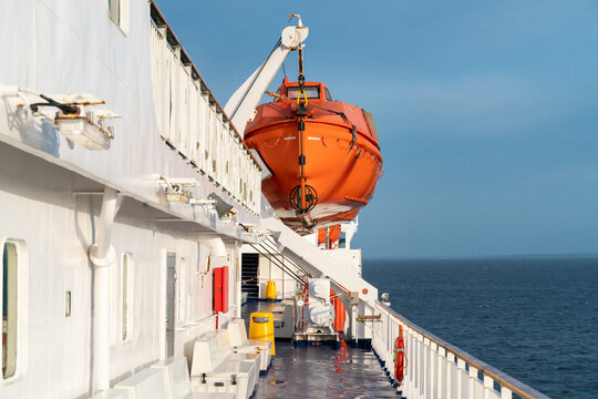 Orange life boat hanging on a crane on a deck of sailing ocean ship with ocean horizon in the back and wooden railing in front.