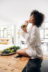Young african american woman drinking green juice with reusable bamboo straw on kitchen counter....
