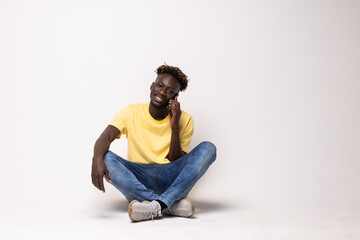 Portrait of a smiling afro american man sitting on the floor talking on the phone isolated on a white background