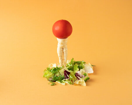 Parsley root like a woman's legs and red tomato standing on mixed salad on yellow background. Minimal food arrangement.