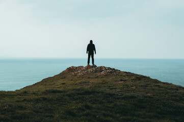 A lone hooded figure standing on top of a hill, looking out across the ocean.