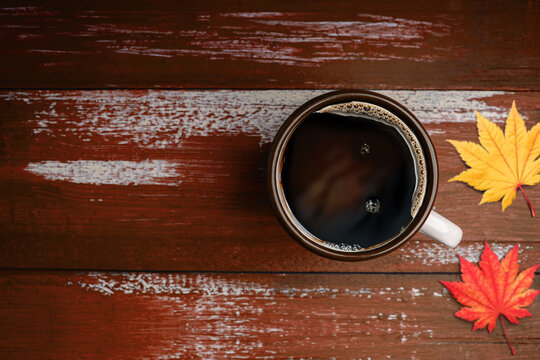 Drinking Coffee in Fall and Autumn Season. Hot Coffee Cup on Orange Brown Wooden Table. Top View. Focus on Cup. surrounded by blurred Maple Leaf