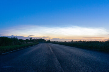 Obraz premium A country road heading into the distance. With the glow of Noctilucent clouds in the morning sky