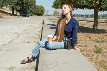 Photo of a beautiful young woman sitting on the side of the embankment with her legs crossed. The...