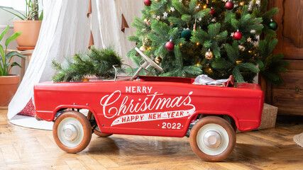 Red retro car near the Christmas tree. A toy car with the inscription "Merry Christmas and Happy New Year 2022" stands near a beautiful Christmas tree in the children's room with a wigwam teepee. © nieriss