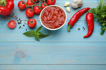 Delicious adjika sauce in bowl and ingredients on light blue wooden table, flat lay. Space for text