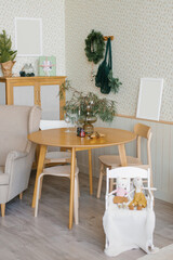 Christmas decor in the living room in the Scandinavian style: two glasses on a wooden table, spruce branches in a vase and candles, a high chair with toys