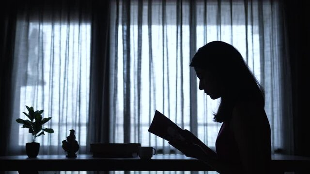 Young Indian Woman reading a book while sitting on a table and flipping its pages at home in evening.A woman  learn and read a  book holding hands closeup with a tea cup and a plant.