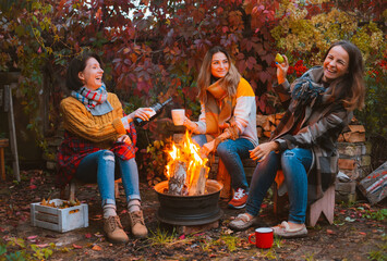 Three happy joyful best friends sitting outside in autumn garden by campfire with bottle of red wine
