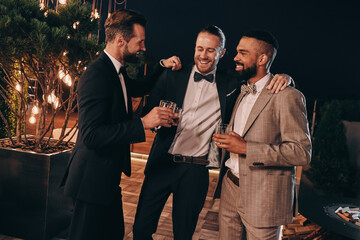 Three handsome men in suits drinking whiskey and communicating while spending time on party