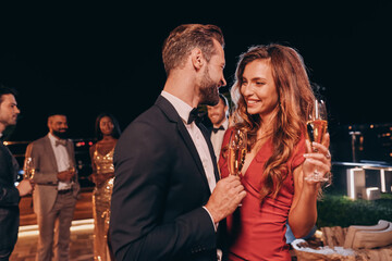 Beautiful young couple in formalwear holding champagne flutes and smiling while spending time on...