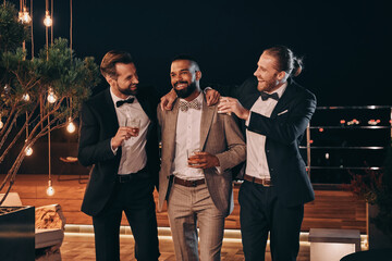 Three handsome men in suits drinking whiskey and communicating while spending time on party