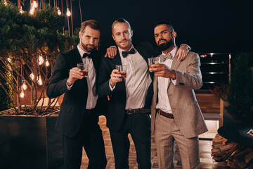 Three cheerful men in suits holding whiskey glasses and looking at camera while spending time on...