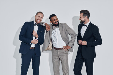 Three handsome young men in suits and bowties looking at camera and smiling while standing against...