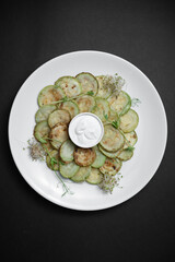 closeup of fried zucchini slices with sour cream on plate on black background