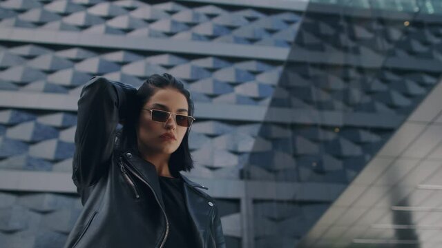 Confident young brunette woman in trendy stylish look wearing black leather jacket and sunglasses looking at camera standing by building in city street, touches her hair. Lifestyle, fashion, urban