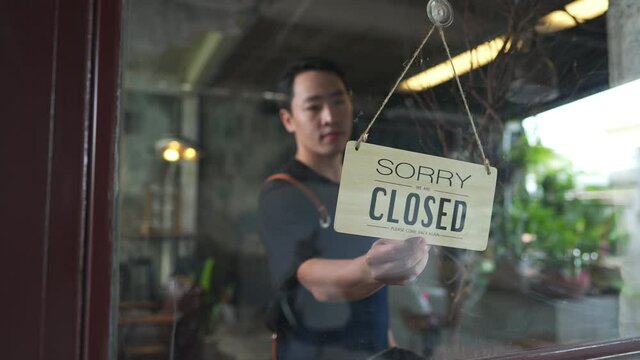 Asian man coffee shop barista walking to cafe door and turning hanging closed sign to open. Male waiter preparing restaurant service to customer. Small business owner and part time job working concept