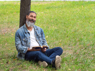Naklejka na ściany i meble Middle aged man with a book in the park. He sits on the grass in the park and looking at camera. Digital detox concept.