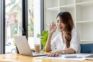 Conceptual Asian businesswoman is video calling with coworker, she is greeting people in chat, she greets with friendly smile, she looks at laptop to chat with friend. Using technology to communicate.