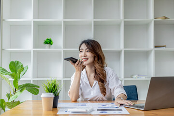 Fototapeta na wymiar Businesswoman talking on the phone with her business partner, she sits in a private room, in a room decorated with plants and bookshelves on the desk with work papers and laptop. Asian women concept.