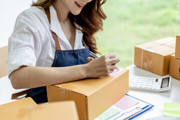 Asian woman writing customer's shipping information on parcel box, she owns an online store, she ships products to customers through a private courier company. Online selling concept.