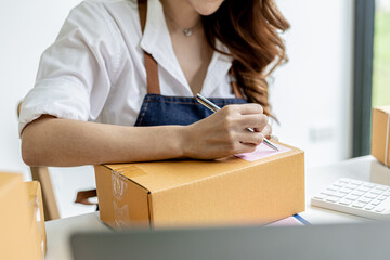 Asian woman writing customer's shipping information on parcel box, she owns an online store, she ships products to customers through a private courier company. Online selling concept.