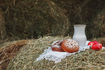 Rustic still life: a jug of milk, red apples and homemade bread on a white tablecloth on a roll of hay. Traditional farm lunch. Food photo.