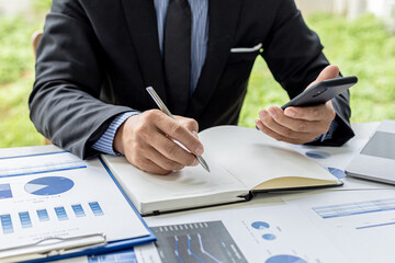 The businessman is taking notes from their mobile phone in a notebook, he owns the company, he is checking company financial documents in his office. Concept of financial management.