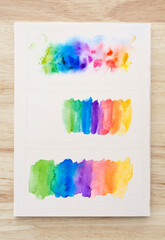 Colorful watercolor brush strokes on white paper sheet with wood background.