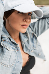 Cool stylish portrait of a beautiful young woman with a sexy look in a fashion blue cap and a blue denim jacket with lace tank top sits on the white sand