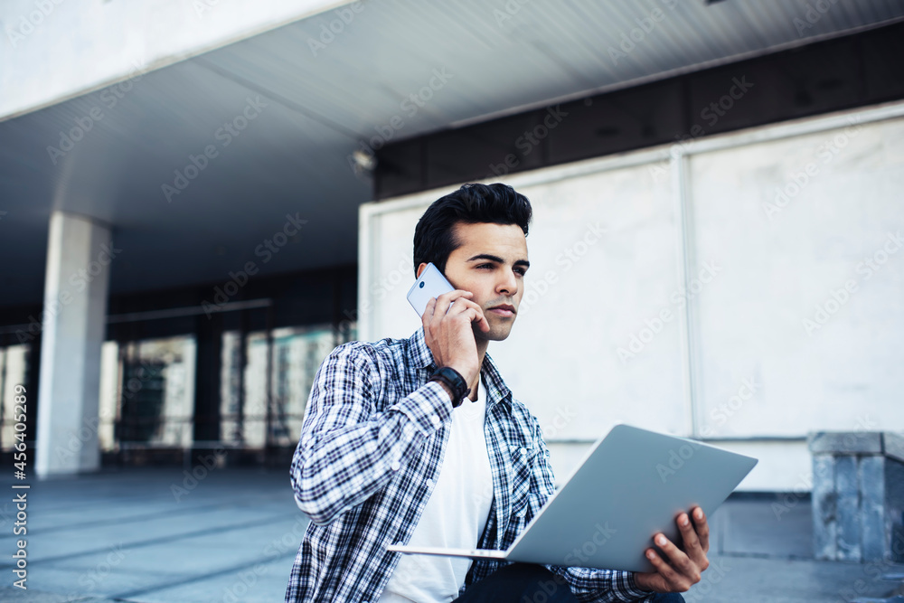 Poster skilled man with modern laptop computer making consultancy cellular conversation during daytime near
