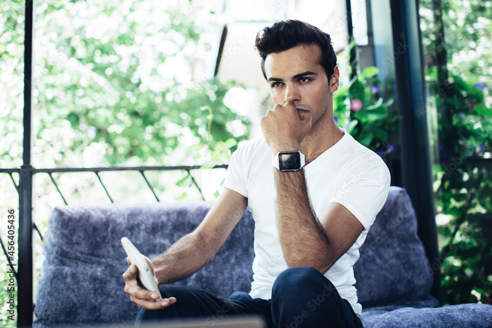 Poster thoughtful male blogger with mobile device pondering on ideas for release text for sharing at web pa