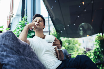 Contemplative Caucaisan male customer visiting cafeteria terrace for drinking tasty caffeine beverage and think about fashionista lifestyle, young hipster guy with coffee cup feeling pondering