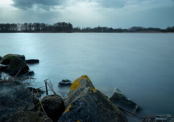 This is a long exposure photo of a lake. On the foreground there are rocks in the water. You also see a tree line and the sky with clouds. The water is smooth because of the long exposure shot.