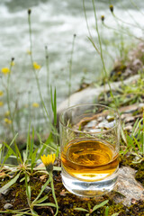 Glass of strong scotch single malt whisky with fast flowing mountain river and wild flowers on background