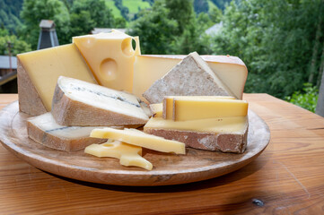 Cheese collection, wooden board with French cheeses comte, beaufort, abondance, emmental, morbier and french mountains village in Haute-Savoie on background