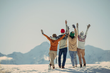 Four happy stylish friends stands, embrace and looks at snow capped mountains. Winter vacations...