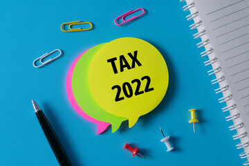 Tax 2022 written in a sticky notes on blue background with pen, notebook, paper clips and push pins.
