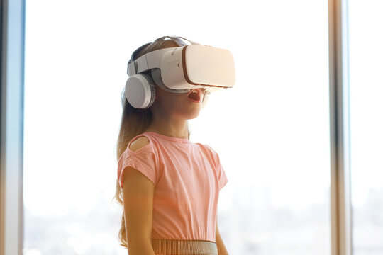 A child is having fun in white virtual reality glasses, standing against the background of a window. High quality photo