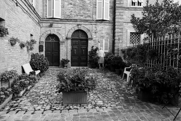 Courtyard of an ancient building in a medieval village in the Marche region, an old woman from behind waters her plants, black and white photo.