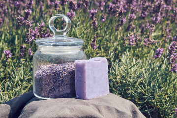 Homemade lavender soap bar and glass jar of dry lavender flowers, blooming lavender field on background.