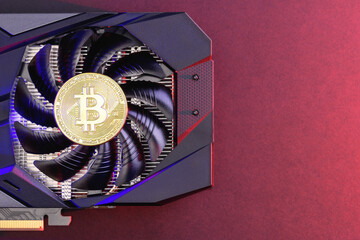 Gold Bitcoin coin on a Black video Card with red and blue backlight, a fan. Crypto currency. Bitcoin mining concept, copy space
