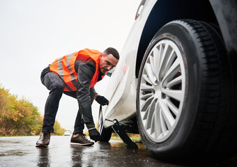 Roadside assistance worker raising vehicle with car jack before changing flat tire. Young man using...