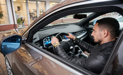 Close-up view of confident, bearded man testing new car in the courtyard of car dealership. Guy putting his hands on the steering wheel and looking straight ahead.
