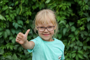 A small blonde girl with glasses gives a thumbs up gesture in a mint T-shirt on a green background,...