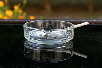 Close up of a  transparent ashtray with a lit cigarette reflected on a black glass table
